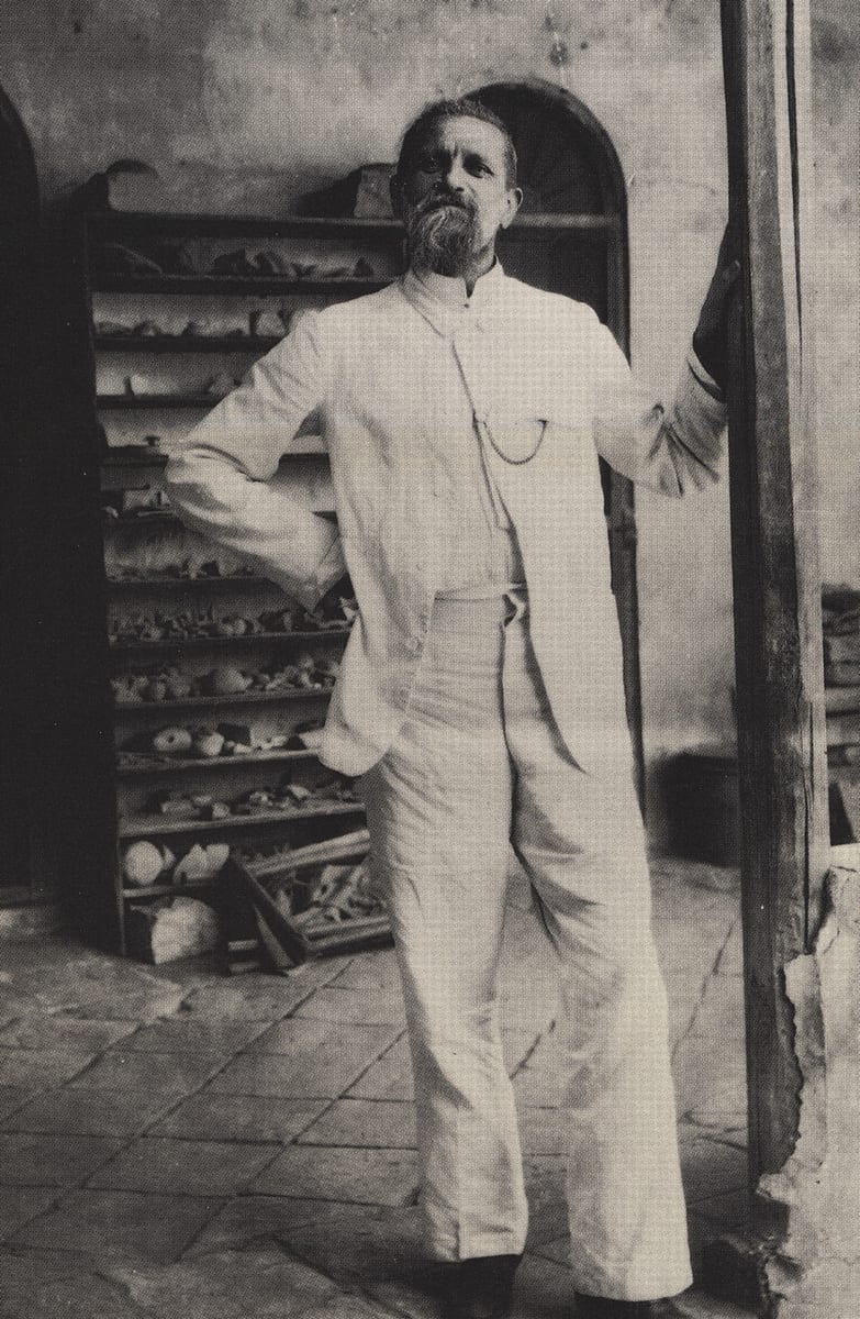 Fotografia antiga de Robert Koldewey, de roupa clara, em pé diante de uma prateleira de peças de cerâmica
