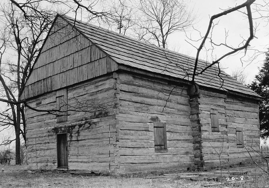 Fotografia em preto e branco do Cane Ridge Meeting House, uma casa de culto de troncos com telhado de tábuas de duas águas, entre árvores sem folhas