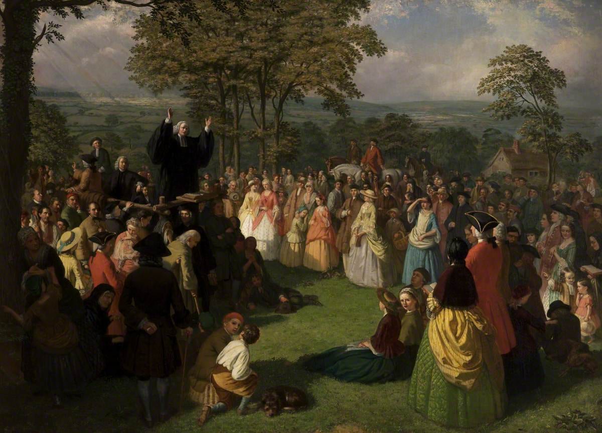 Pintura de George Whitefield de beca preta e braços erguidos, pregando de um púlpito improvisado a uma grande multidão ao ar livre, sob árvores
