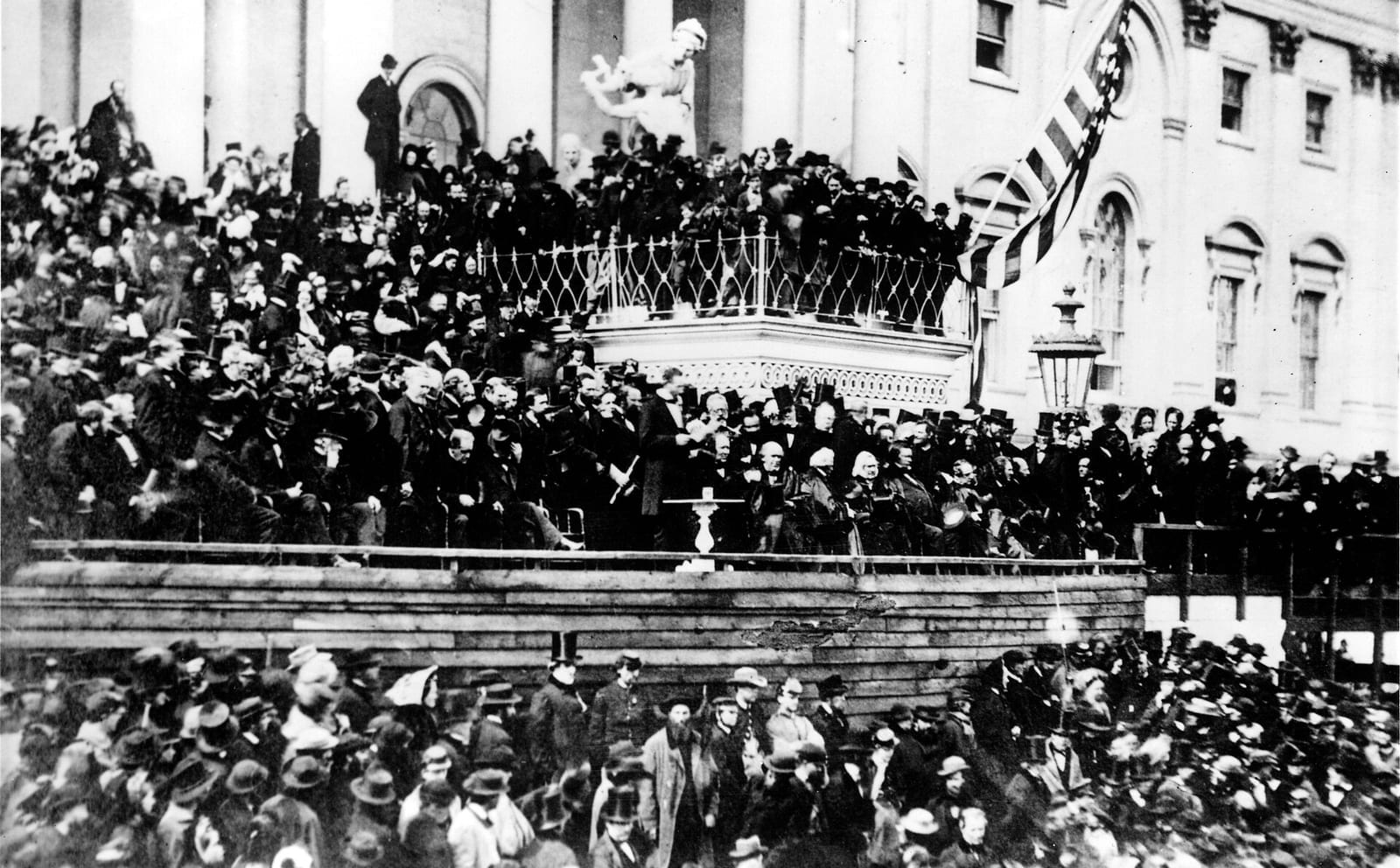Fotografia antiga do Segundo Discurso de Posse de Lincoln em frente ao Capitólio: o presidente de pé lendo, cercado de homens de casaca, com grande multidão e uma bandeira
