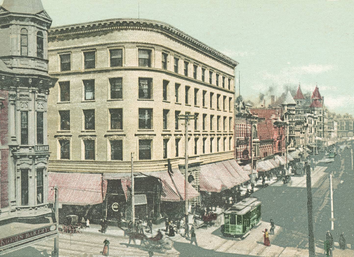 Cartão-postal colorido de uma rua movimentada de Los Angeles por volta de 1903, com um bonde elétrico verde, carroças puxadas por cavalos, pedestres, toldos listrados e um prédio de esquina de vários andares