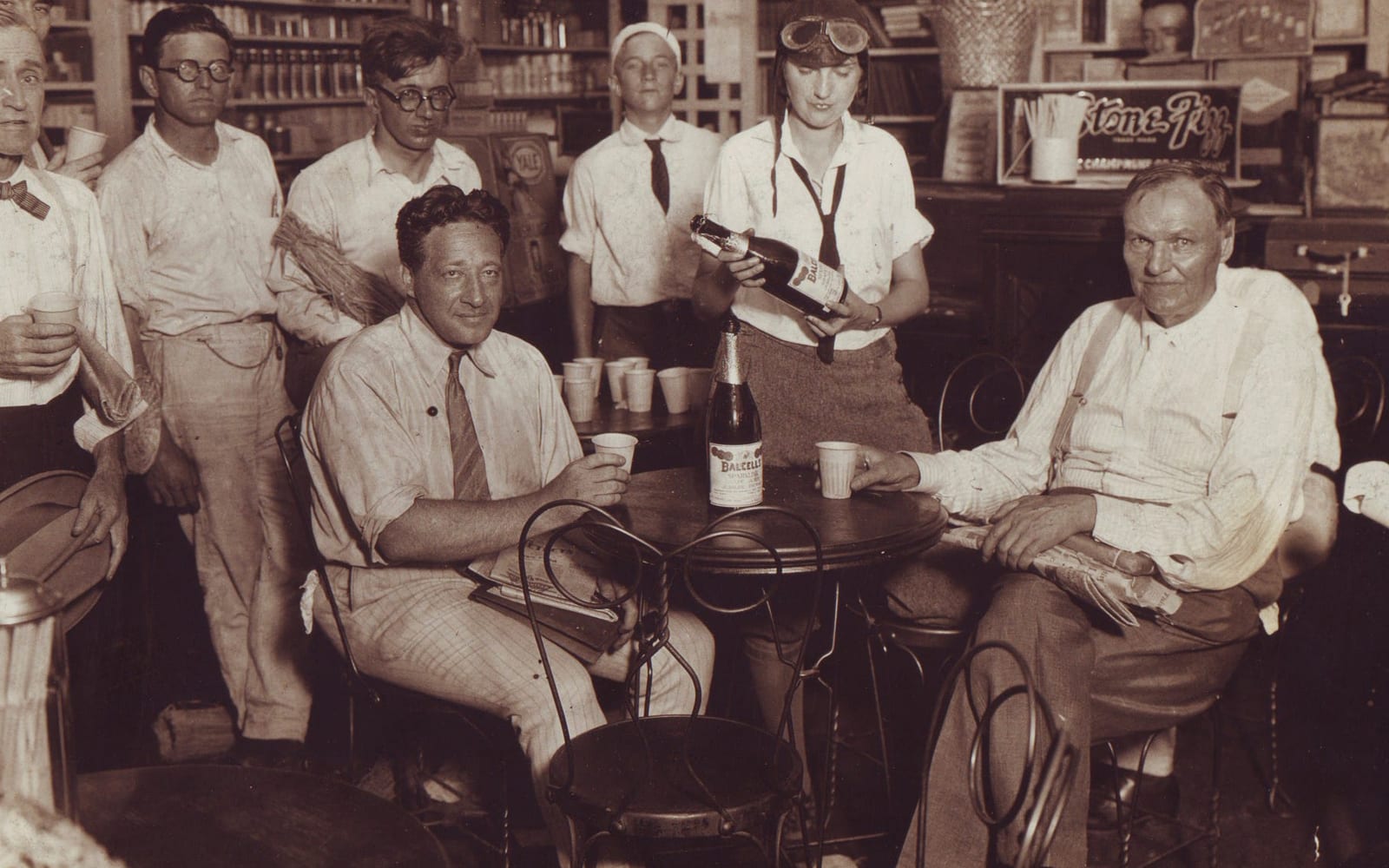 Fotografia antiga em tons de sépia, dentro de uma loja de remédios de cidade pequena: dois homens sentados a uma mesa redonda com copos e uma garrafa de suco de uva, e mulheres e homens de pé ao fundo, diante de prateleiras