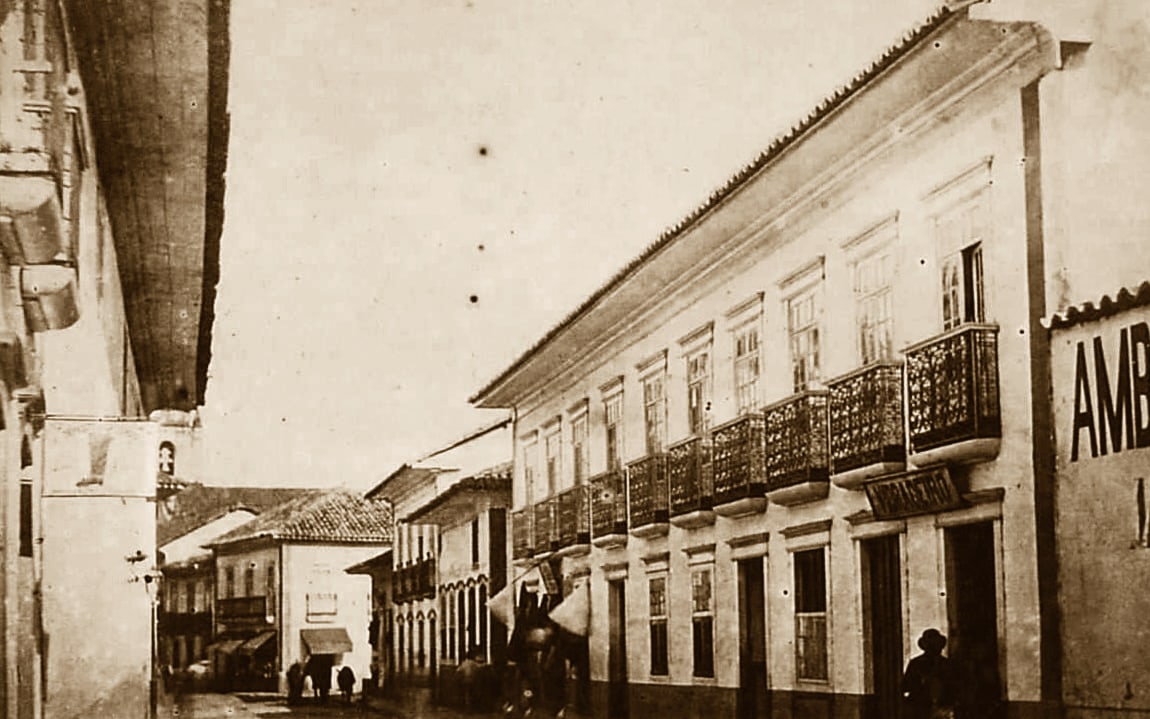 Fotografia em tons de sépia de uma rua estreita de casarões de dois andares com balcões de ferro, num dia sem trânsito, com poucas pessoas ao fundo