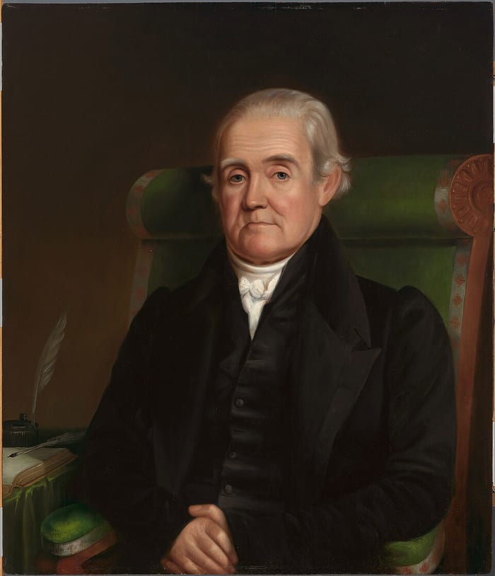 Retrato a óleo de Noah Webster idoso, de cabelo branco e casaco escuro, sentado numa poltrona verde, com uma pena e um livro aberto sobre a mesa ao lado