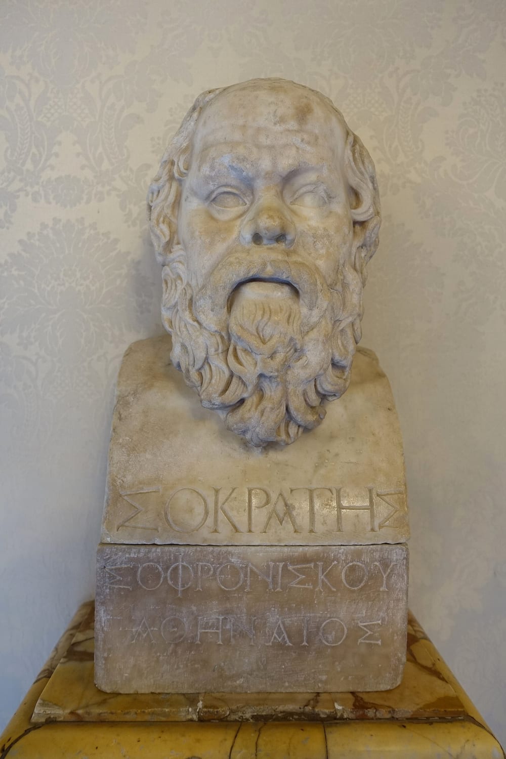 Busto de mármore de Sócrates: rosto largo, calvo na testa, barba cheia e olhos fundos, sobre um pilar com inscrição grega