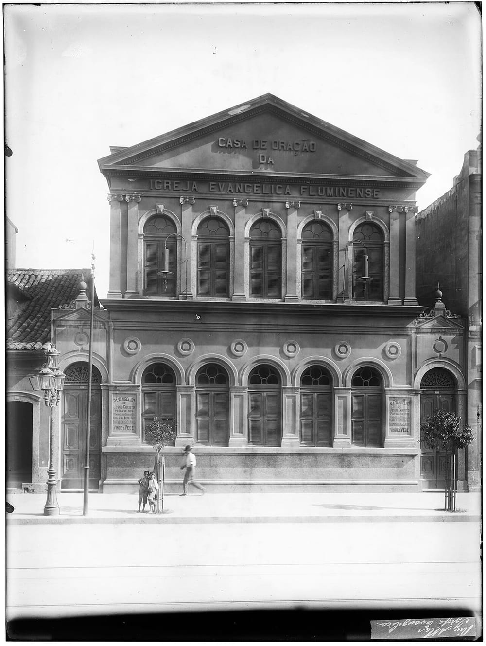 Fotografia antiga em preto e branco da fachada de um edifício de dois andares com frontão, onde se lê Casa de Oração da Igreja Evangélica Fluminense; painéis nas portas laterais convidam ao culto; duas crianças e um homem na calçada