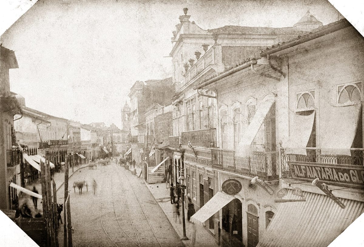 Fotografia antiga em sépia de uma rua de São Paulo com sobrados de balcões de ferro, trilhos de bonde no meio do calçamento, toldos e uma placa de alfaiataria; ao fundo, as torres de uma igreja