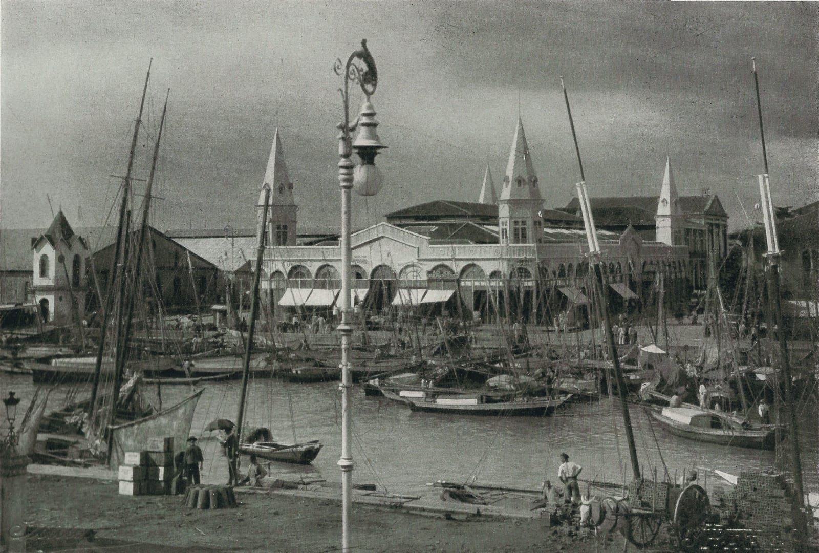 Fotografia em preto e branco do mercado de ferro do Ver-o-Peso, em Belém, com torres pontiagudas ao fundo, muitos barcos a vela e canoas atracados na doca e um poste de luz em primeiro plano