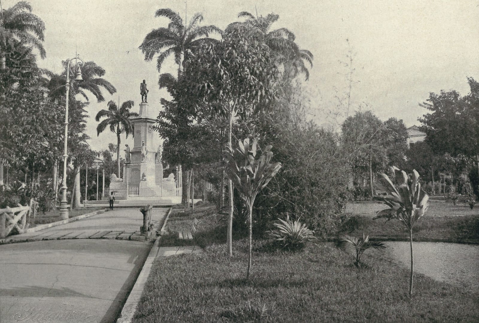 Fotografia antiga em preto e branco de uma praça de Belém: caminho de pedra à esquerda, palmeiras altas e um monumento com estátua ao fundo, canteiros de plantas em primeiro plano